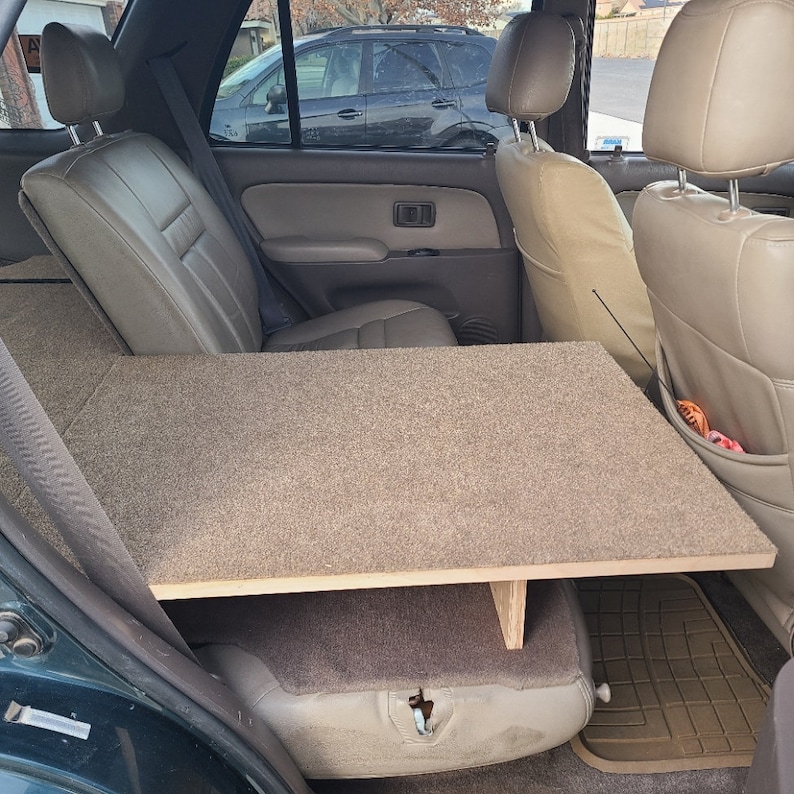May include: A green SUV with the back seats folded down and a wooden platform covered in brown carpet installed to create a flat sleeping surface.