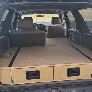 May include: A black SUV with the back seats folded down and a custom built wooden platform with two drawers in the cargo area. The platform is covered in beige carpet.
