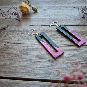 Puede incluir: Un par de pendientes colgantes con forma rectangular. Los pendientes están hechos de madera y pintados en un esquema de colores verde oscuro y rosa. Los pendientes tienen un gancho plateado.