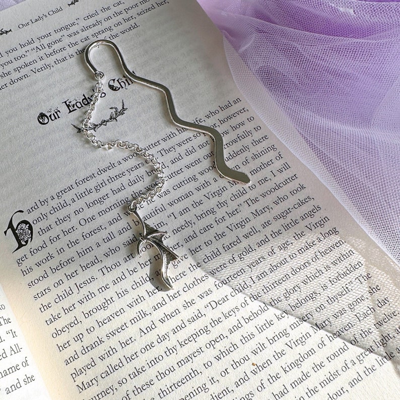 May include: A silver bookmark shaped like a tree branch with a chain attached. The bookmark is placed on an open book with text about a woman named Mary. The text reads 'Our Lady's Child' and 'Our Lady Child'.