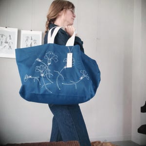 May include: A large, blue tote bag with white floral embroidery. The bag has white handles and a tag attached. The design features delicate white floral stems and blooms across the front of the bag. The bag is made of a sturdy fabric.