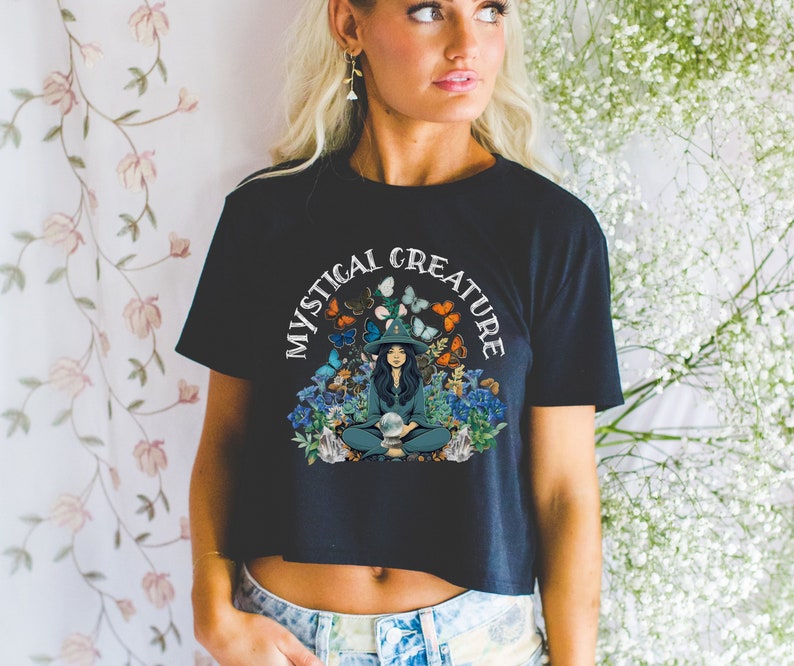 Może przedstawiać: Czarny crop top z nadrukiem kobiety w kapeluszu czarownicy siedzącej z kryształową kulą otoczoną motylami i kwiatami. Tekst "Mystical Creature" jest napisany w k&oacute;łku wok&oacute;ł nadruku.