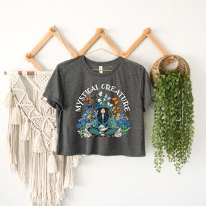 Może przedstawiać: Kr&oacute;tki t-shirt w kolorze ciemnoszarym z grafiką przedstawiającą kobietę w kapeluszu czarownicy otoczoną motylami i kwiatami. Wok&oacute;ł grafiki widnieje napis "Mystical Creature".