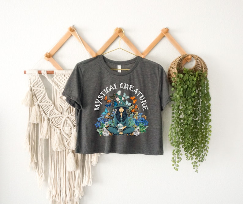 Może przedstawiać: Kr&oacute;tki t-shirt w kolorze ciemnoszarym z grafiką przedstawiającą kobietę w kapeluszu czarownicy otoczoną motylami i kwiatami. Wok&oacute;ł grafiki widnieje napis "Mystical Creature".