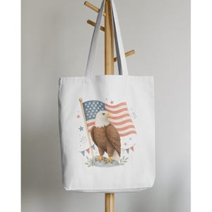 May include: White tote bag featuring a watercolor illustration of an American bald eagle standing before a waving American flag. The flag has red and white stripes and a blue field with white stars. The eagle is brown and white.