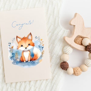 May include: A congratulatory card with a watercolor illustration of a fox in a forest setting. The card has the word "Congrats!" written on it in blue lettering. The card is next to a wooden rocking horse toy with a beaded teething ring.