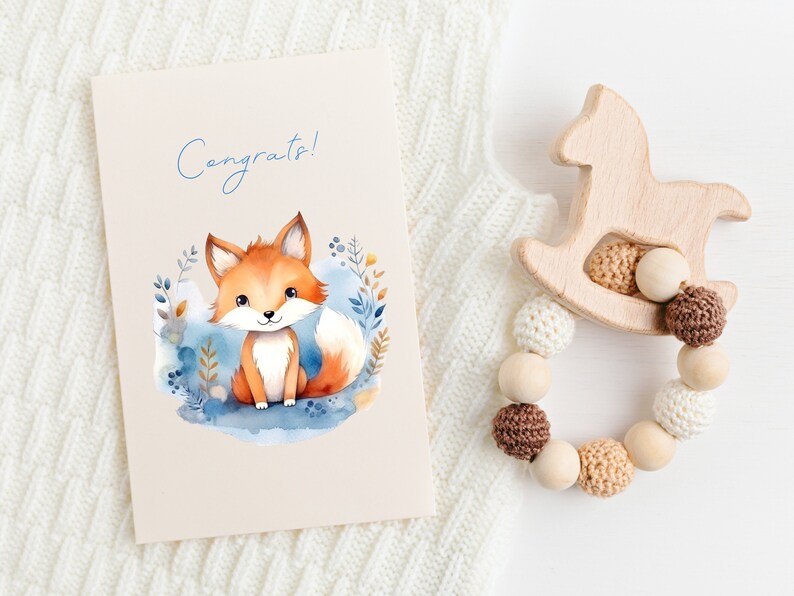 May include: A congratulatory card with a watercolor illustration of a fox in a forest setting. The card has the word "Congrats!" written on it in blue lettering. The card is next to a wooden rocking horse toy with a beaded teething ring.