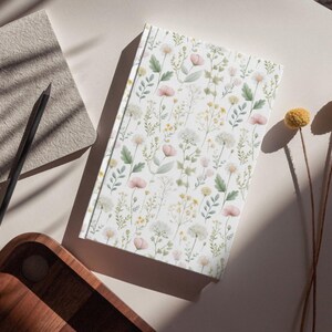 May include: A floral-patterned journal with a white cover featuring delicate pink, yellow, and green botanical illustrations. A black pencil and a textured notepad sit nearby. Dried yellow flowers add a natural touch to the scene.
