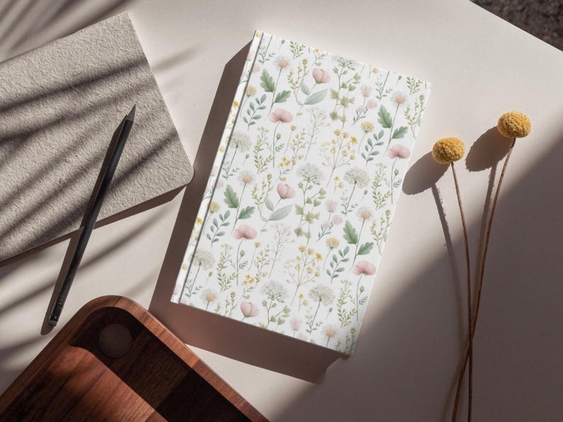 May include: A floral-patterned journal with a white cover featuring delicate pink, yellow, and green botanical illustrations. A black pencil and a textured notepad sit nearby. Dried yellow flowers add a natural touch to the scene.