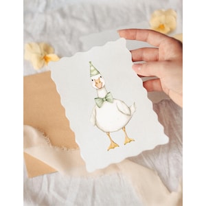 Puede incluir: Ilustraci&oacute;n en acuarela de un ganso blanco con pajarita y gorro de fiesta verdes. El ganso est&aacute; sobre una tarjeta con bordes festoneados, sostenida por una mano. Fondo suave y neutro con flores y una cinta.