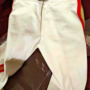 Può includere: Pantaloni bianchi con una striscia rossa e dorata sul lato. I pantaloni hanno una cintura con una fibbia argentata.