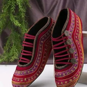 Pode incluir: Um par de sapatos bordados com uma rica paleta de cores. Os sapatos são predominantemente vermelhos, adornados com bordados dourados, roxos e laranja. Têm atacadores vermelho escuro e botões decorativos. O design sugere uma influência cultural ou étnica.