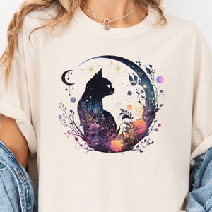 May include: A cream colored t-shirt with a black cat silhouette sitting on a crescent moon. The moon is surrounded by flowers and stars in shades of purple, blue, and orange.
