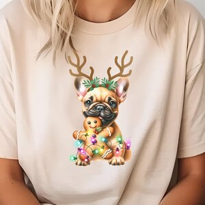 May include: A beige t-shirt with a graphic of a French bulldog wearing reindeer antlers and a Christmas wreath. The bulldog is holding a gingerbread man and surrounded by colorful Christmas lights.
