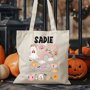 May include: Cream-colored tote bag with the name "Sadie" and the word "Boo!" in black and pink. The bag features a colorful Halloween-themed design with pumpkins, a ghost, a rainbow, and a witch's hat.