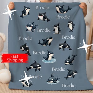 May include: A blue blanket featuring cartoon orcas in black and white, with the name "Brodie" printed in white. The blanket is held up by a person. The words "Fast Shipping" are on a red banner.