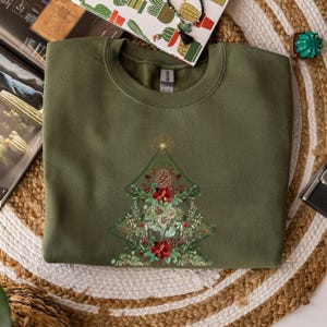 May include: An olive green sweatshirt featuring a detailed Christmas tree design. The tree is composed of pinecones, red flowers, and greenery, with a gold star at the top. The sweatshirt is folded on a woven mat.
