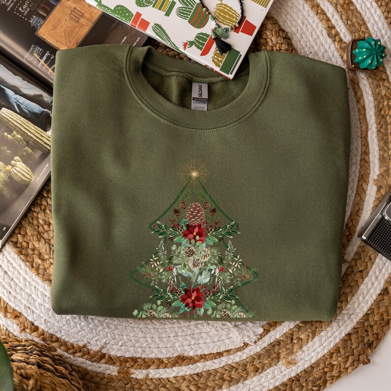 May include: An olive green sweatshirt featuring a detailed Christmas tree design. The tree is composed of pinecones, red flowers, and greenery, with a gold star at the top. The sweatshirt is folded on a woven mat.