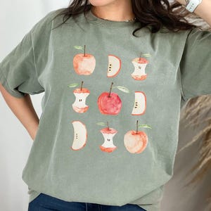 May include: Sage green t-shirt featuring a watercolor design of apples and apple cores. The design includes whole apples, apple slices, and cores in shades of red, pink, and peach. The shirt is a relaxed fit.