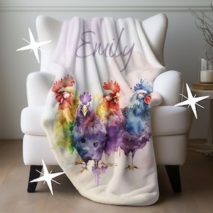 May include: A white plush blanket draped over a white armchair. The blanket features a watercolor design of four colorful roosters and the name "Emily" in script. The roosters are in shades of red, yellow, purple, and blue.