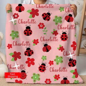 May include: Pink baby blanket with a repeating pattern of red ladybugs, red flowers, and green flowers. The name "Charlotte" is written in red script throughout the design. A white starburst is also included in the design.