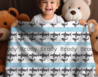 May include: White baby blanket with a blue and gray shark pattern. The blanket has the name "Brody" repeated across the bottom.