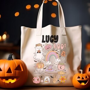 May include: Off-white tote bag with the name "LUCY" and Halloween-themed illustrations. The design includes a ghost, pumpkins, a rainbow, and the word "BOO!". Two carved orange pumpkins flank the bag.
