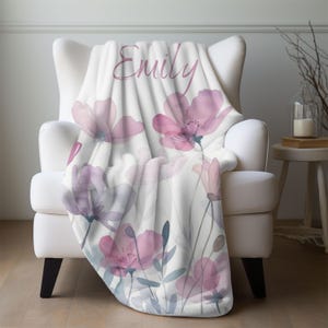 Puede incluir: Una manta de forro polar suave y blanca con un dise&ntilde;o floral de acuarela en tonos rosa y morado. La manta lleva el nombre "Emily" en una fuente de letra cursiva rosa.