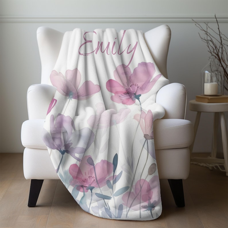 Puede incluir: Una manta de forro polar suave y blanca con un dise&ntilde;o floral de acuarela en tonos rosa y morado. La manta lleva el nombre "Emily" en una fuente de letra cursiva rosa.
