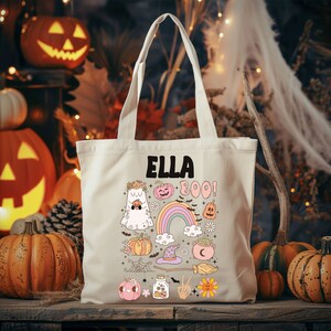 May include: Beige canvas tote bag with the name "ELLA" in black block letters. The bag features a colorful Halloween-themed design with a ghost, pumpkins, rainbow, and the word "BOO!". The bag has two handles.
