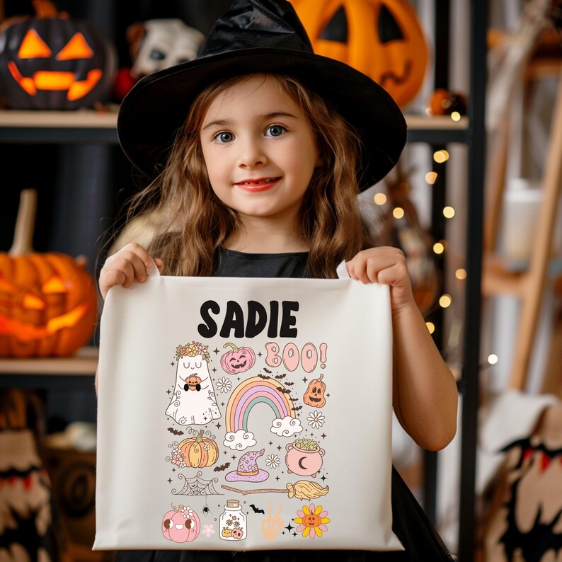 May include: A young girl wearing a black witch hat holds a cream-colored Halloween tote bag. The bag features the name "Sadie" above a collection of Halloween-themed illustrations, including a ghost, pumpkins, a rainbow, and the word "BOO!"