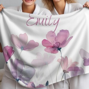 Puede incluir: Una manta blanca con un dise&ntilde;o floral con flores de acuarela rosa y p&uacute;rpura. La manta tiene el nombre "Emily" escrito en letras cursivas rosas.