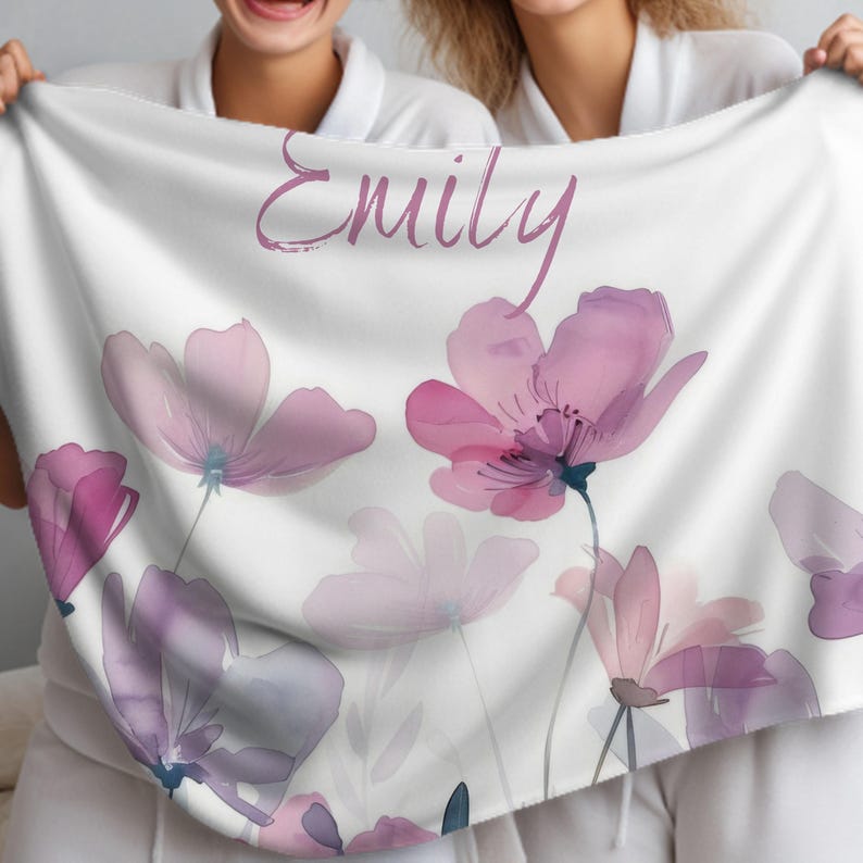 Puede incluir: Una manta blanca con un dise&ntilde;o floral con flores de acuarela rosa y p&uacute;rpura. La manta tiene el nombre "Emily" escrito en letras cursivas rosas.