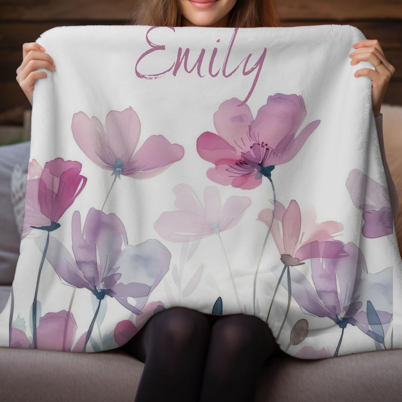 Puede incluir: Una manta de forro polar blanca con un dise&ntilde;o floral de acuarela en tonos rosa y morado. La manta tiene el nombre "Emily" escrito en cursiva.