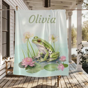 May include: A light blue towel with the name "Olivia" in green script. The towel features a watercolor illustration of a green frog, pink water lilies, and white flowers.