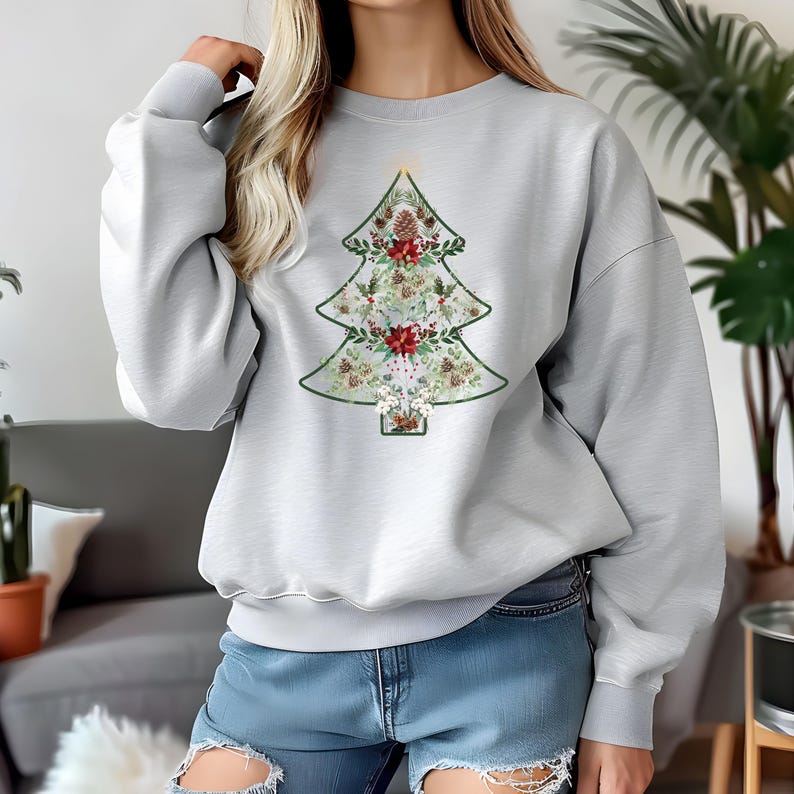 May include: A light gray sweatshirt featuring a Christmas tree design. The tree is outlined in green and filled with floral and pinecone elements in red, white, and brown. The sweatshirt has long sleeves and a crew neck.