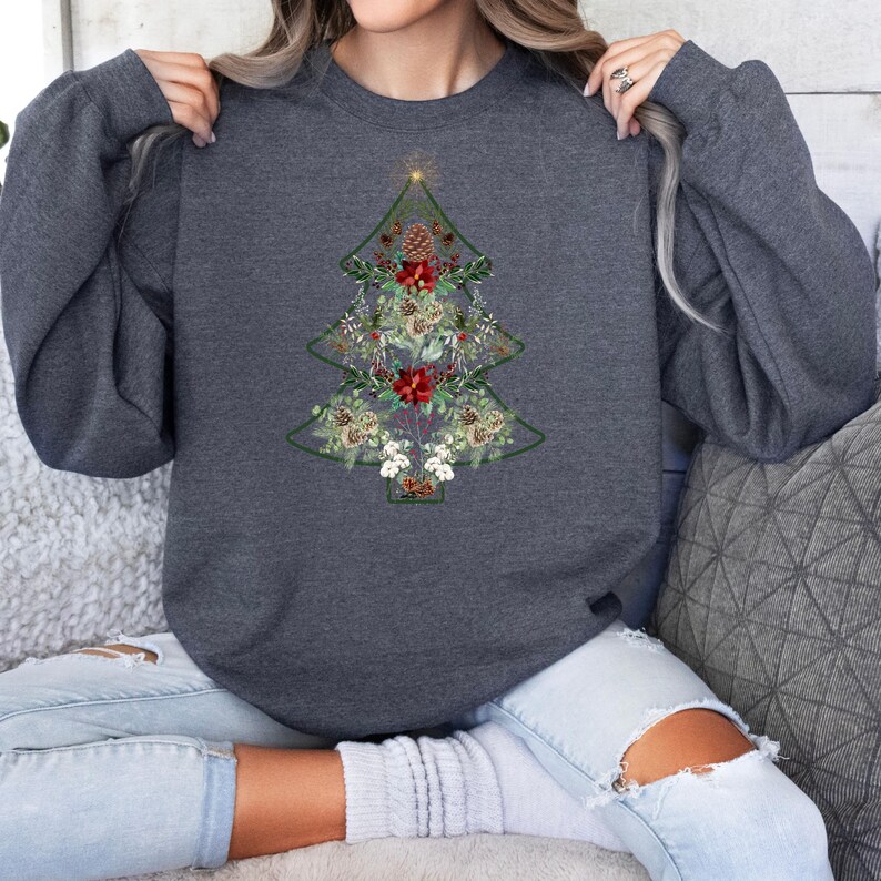May include: A dark gray sweatshirt featuring a Christmas tree design. The tree is outlined in green and filled with floral and pinecone elements in shades of red, white, and beige. The sweatshirt has long sleeves.