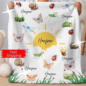 May include: White baby blanket featuring watercolor illustrations of insects, butterflies, and a sun. The name "Harper" is written in script font throughout the design. The blanket has a green grass border.