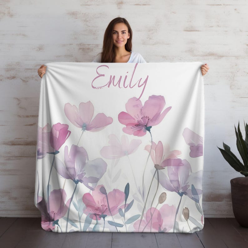 Puede incluir: Una manta de forro polar blanca con un dise&ntilde;o floral de acuarela en tonos rosa y morado. La manta tiene el nombre "Emily" escrito en cursiva rosa.