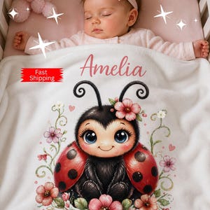 May include: A white baby blanket featuring a watercolor illustration of a cute ladybug with big blue eyes, red wings with black spots, and pink flowers. The name "Amelia" is printed in pink.