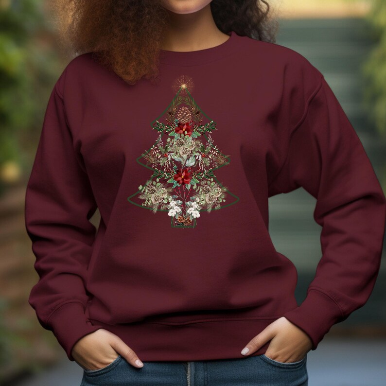 May include: Burgundy sweatshirt featuring a Christmas tree design. The tree is composed of floral and botanical elements, including red poinsettias, pinecones, and greenery. A gold star tops the tree. The sweatshirt has a crew neck and long sleeves.