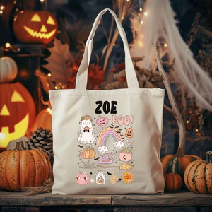 May include: Beige tote bag with a Halloween-themed design. The design includes a ghost, pumpkins, a rainbow, and the text "Zoe Boo!". The bag has two handles and is sitting on a wooden surface with pumpkins in the background.