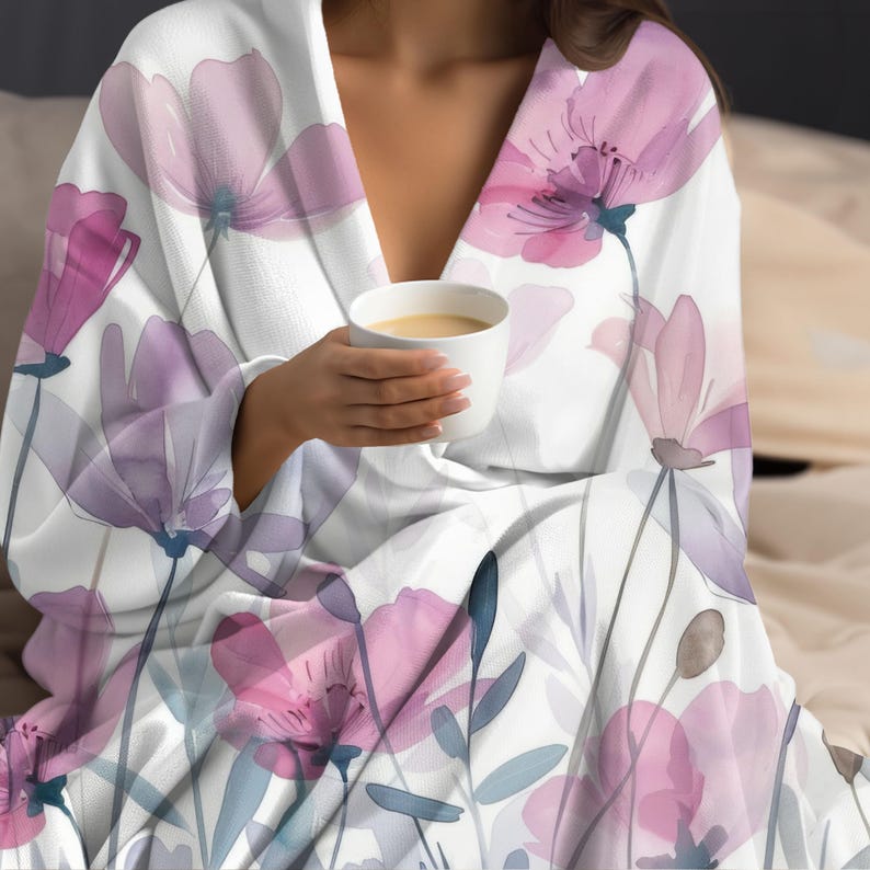 Puede incluir: Una manta blanca con un patr&oacute;n floral de acuarela en tonos de rosa y morado. La manta est&aacute; drapeada sobre una persona que sostiene una taza de caf&eacute;.
