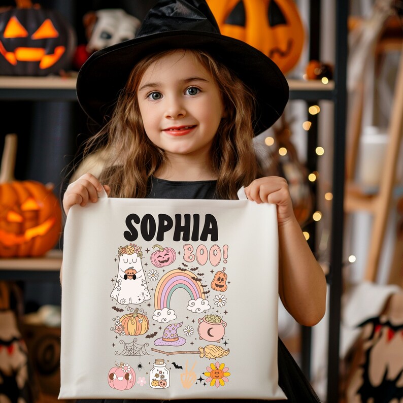 May include: A young girl wearing a black witch hat holds a cream-colored tote bag. The bag features the name "SOPHIA" above a collection of Halloween-themed illustrations, including a ghost, pumpkins, a rainbow, and the word "BOO!"