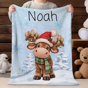 May include: A light blue fleece blanket featuring the name Noah in black text. The design includes a cartoon moose wearing a red Santa hat and a green striped scarf, surrounded by white snowy pine trees and light blue snowflakes.