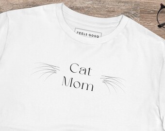May include: White t-shirt with the text "Cat Mom" and a stylized cat whisker design. The shirt is laid out on a wooden surface with a pair of black sneakers and blue jeans.