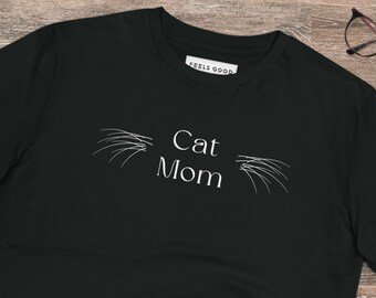 May include: Black t-shirt with white text that reads "Cat Mom" and stylized cat whiskers above the text. The shirt is laying on a wood surface with a pair of jeans and sneakers.