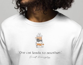 Op de afbeelding: Wit sweatshirt met een zwart-witte illustratie van een stapel katten. De tekst "'One cat leads to another.'" is onder de illustratie gedrukt, met de naam "Ernest Hemingway" onder de tekst.