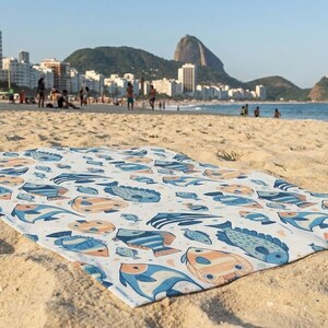 A colorful beach towel with fish patterns is spread out on a sandy beach, with a city skyline and mountains visible in the background.