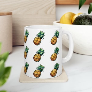 A white mug with a repeating pattern of pineapples on a wooden coaster, placed on a white countertop.